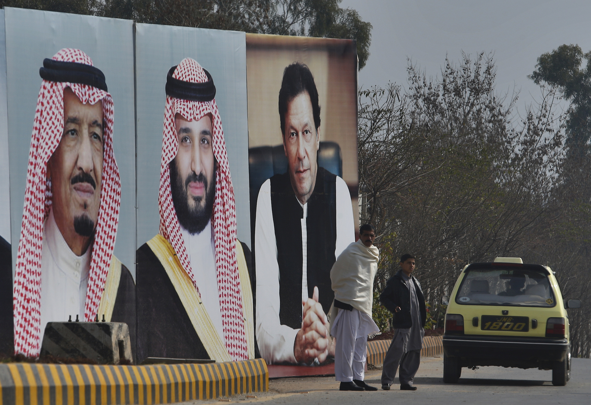 A man with his son waits next to portraits of Saudi leaders and a Pakistani prime minister displayed on a highway on the occasion of the visit by Saudi Arabia's crown prince to Pakistan, in Islamabad, Pakistan, Feb. 17, 2019. (AP Photo/Anjum Naveed)