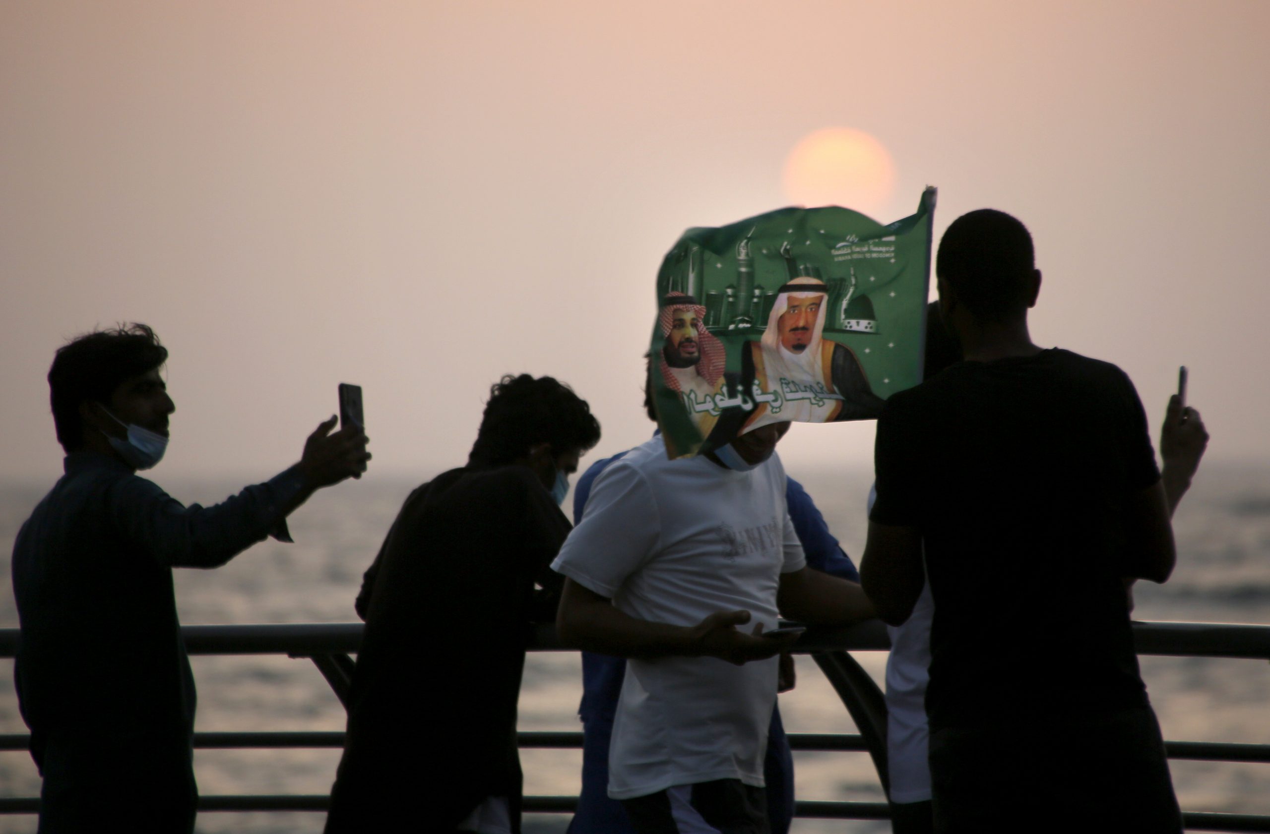 Saudis watch the sunset as they wave a flag with pictures of Saudi King Salman bin Abdulaziz, right, and Crown Prince Mohammed bin Salman during a celebration marking National Day, in Jeddah, Saudi Arabia, Sept. 23, 2020. (AP Photo/Amr Nabil)