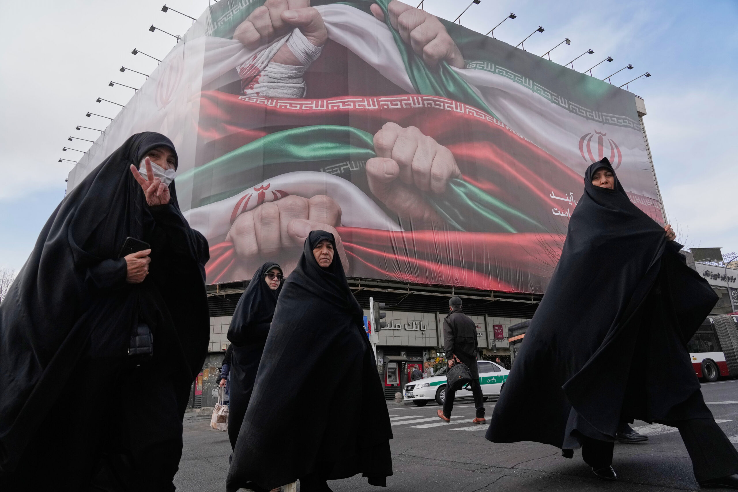 Women cross a street under a banner showing hands firmly holding Iranian flags as a sign of patriotism, as one of them flashes the victory sign, in Tehran, Iran, Jan. 14. (AP Photo/Vahid Salemi)
